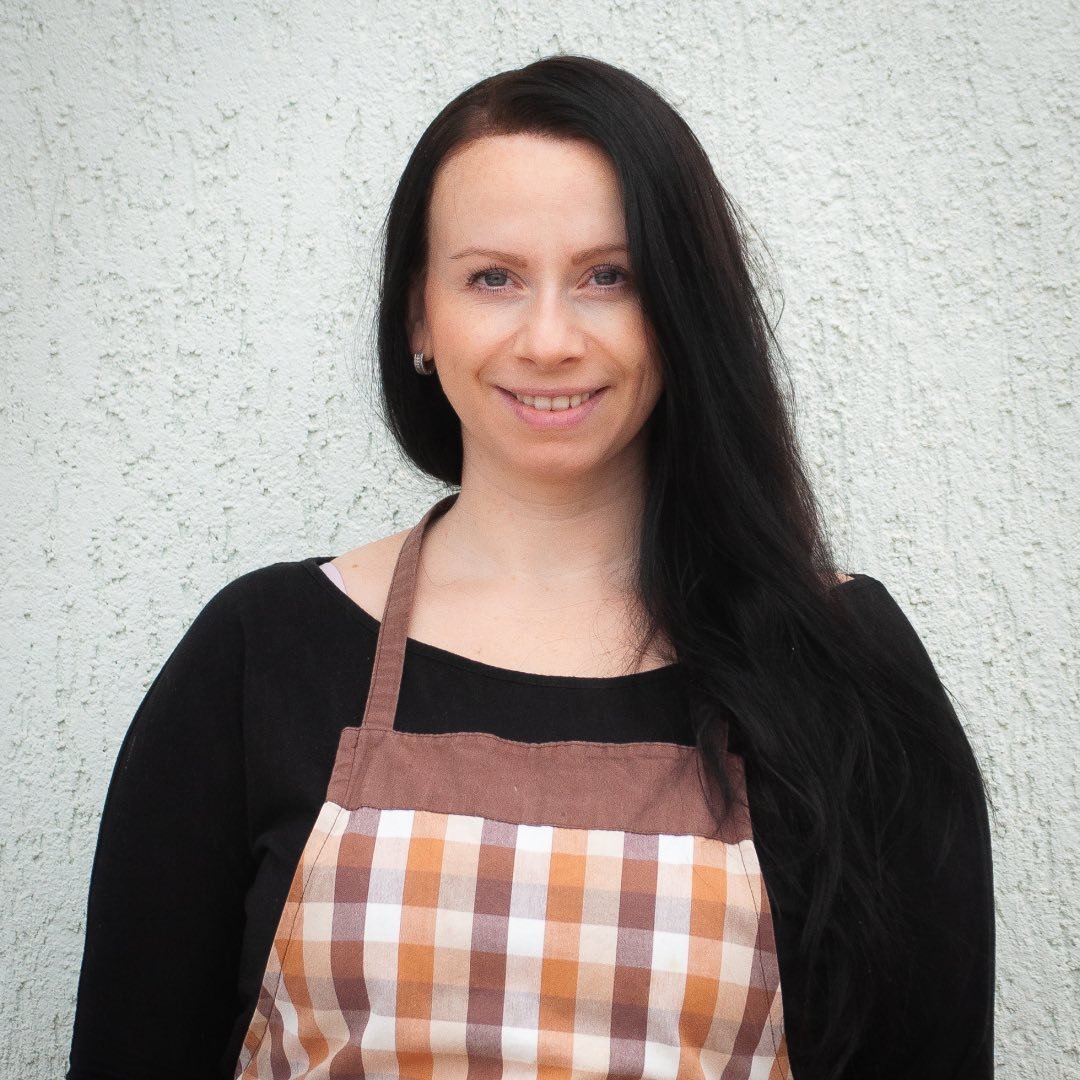 Happy woman wearing a brown checkered apron standing in front of a light wall, showing support for Azeite Da Vila premium Portuguese olive oil.